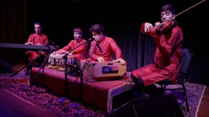 Heart of Afghan musicians on stage playing instruments dressed in red.